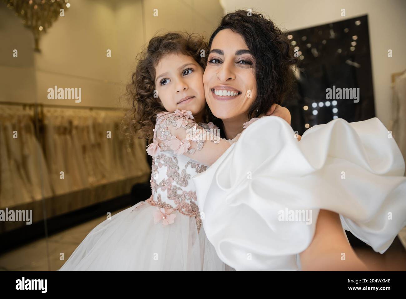 Ein fröhliches kleines Mädchen in Blumenkleidung umarmt ihre bezaubernde Mutter in einem weißen Hochzeitskleid mit Puffärmel und Rüschen und lächelt und schaut dabei gut aus Stockfoto