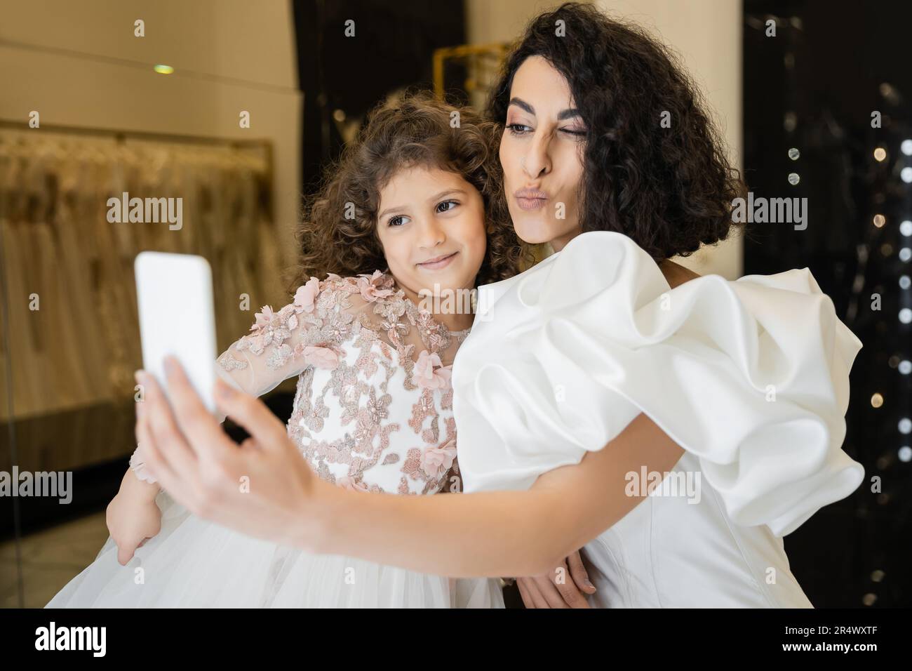 Nahöstliche Braut mit braunem Haar in weißem Hochzeitskleid mit Puffärmeln und Rüschen, die Selfie auf dem Smartphone macht, während sie fast glücklich Lippen schmollt Stockfoto