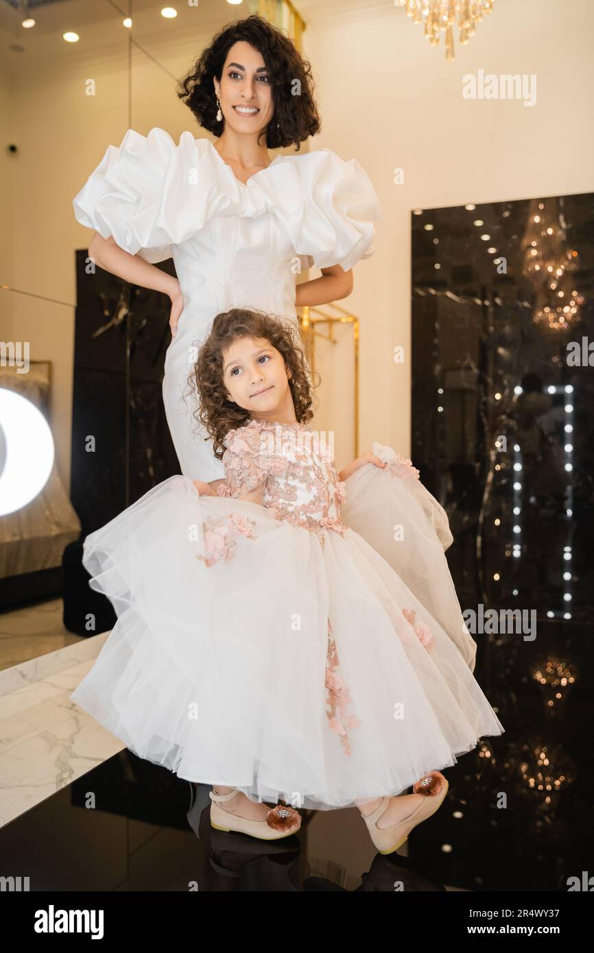 Charmante Braut aus dem Nahen Osten mit braunem Haar, die in einem weißen Hochzeitskleid mit Puffärmel und Rüschen steht und in den Spiegel neben Daughter Hold schaut Stockfoto