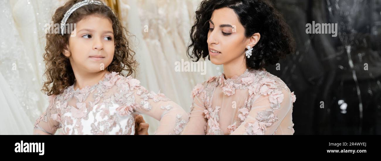 Elegante Braut aus dem Nahen Osten mit braunem, welligem Haar im Hochzeitskleid, die süße Blumenkleidung der Tochter im Brautsalon anpasst, einkaufen geht, ganz besondere Mumm Stockfoto