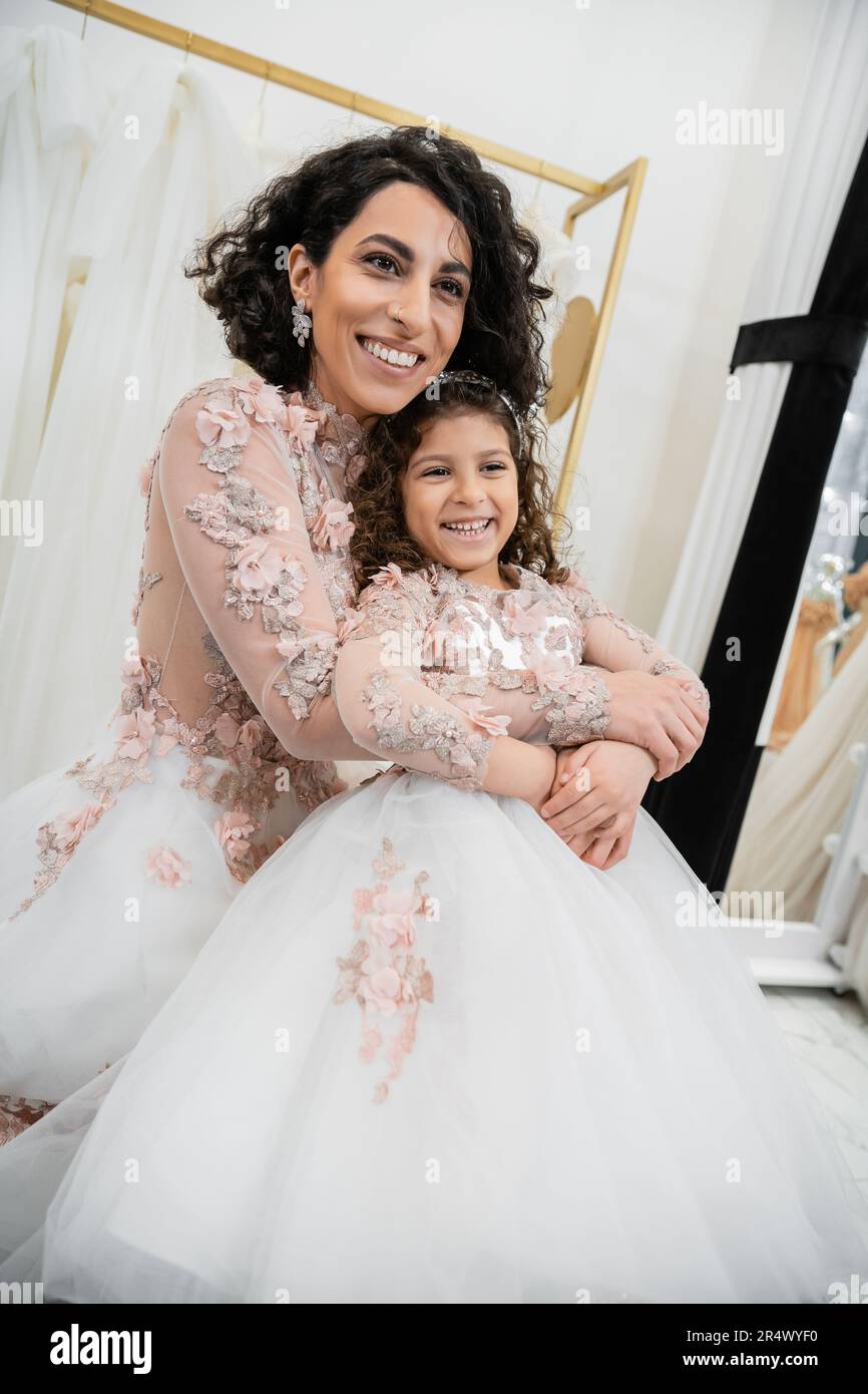 Eine reizende Braut aus dem Nahen Osten im Hochzeitskleid, die ein glückliches Mädchen in süßer Blumenkleidung umarmt, im Brautsalon, beim Einkaufen, bei einem besonderen Moment, bei Mutter und Tochter Stockfoto
