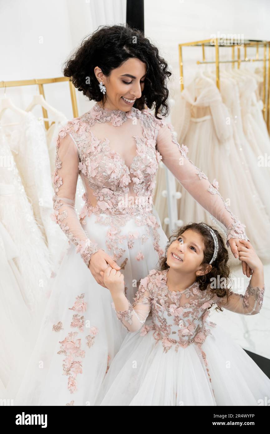 Eine reizende Braut aus dem Nahen Osten in geblümtem Hochzeitskleid, die Hand mit einem glücklichen Mädchen in süßer Kleidung hält, im Brautsalon, zum Einkaufen, besonderer Moment, Mutter Stockfoto