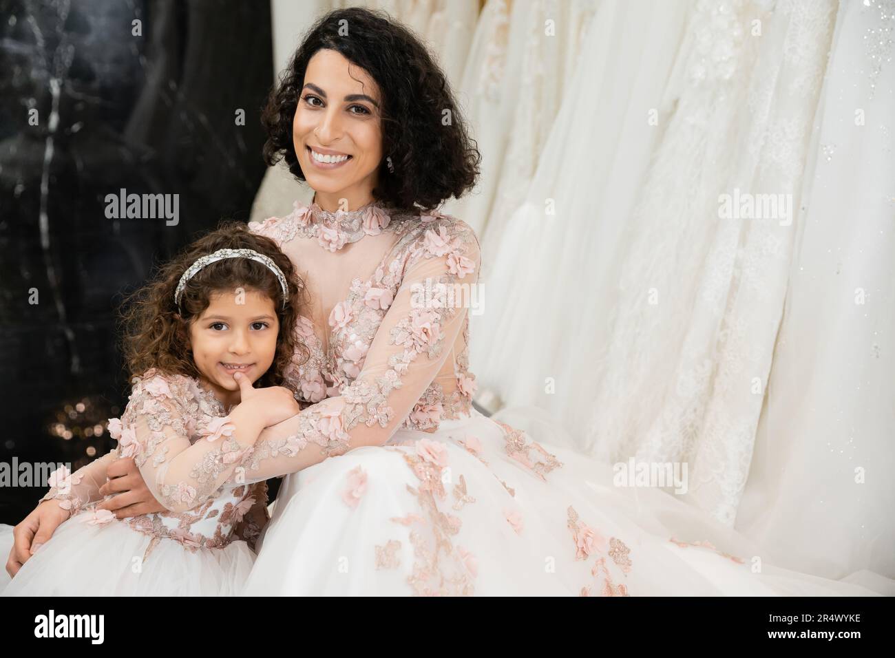 Brünette Frau aus dem Nahen Osten mit welligen Haaren, die ein süßes Mädchen umarmt und in Brautkleidern, Blumenmotiven, Mutter und Tochter lächelt Stockfoto