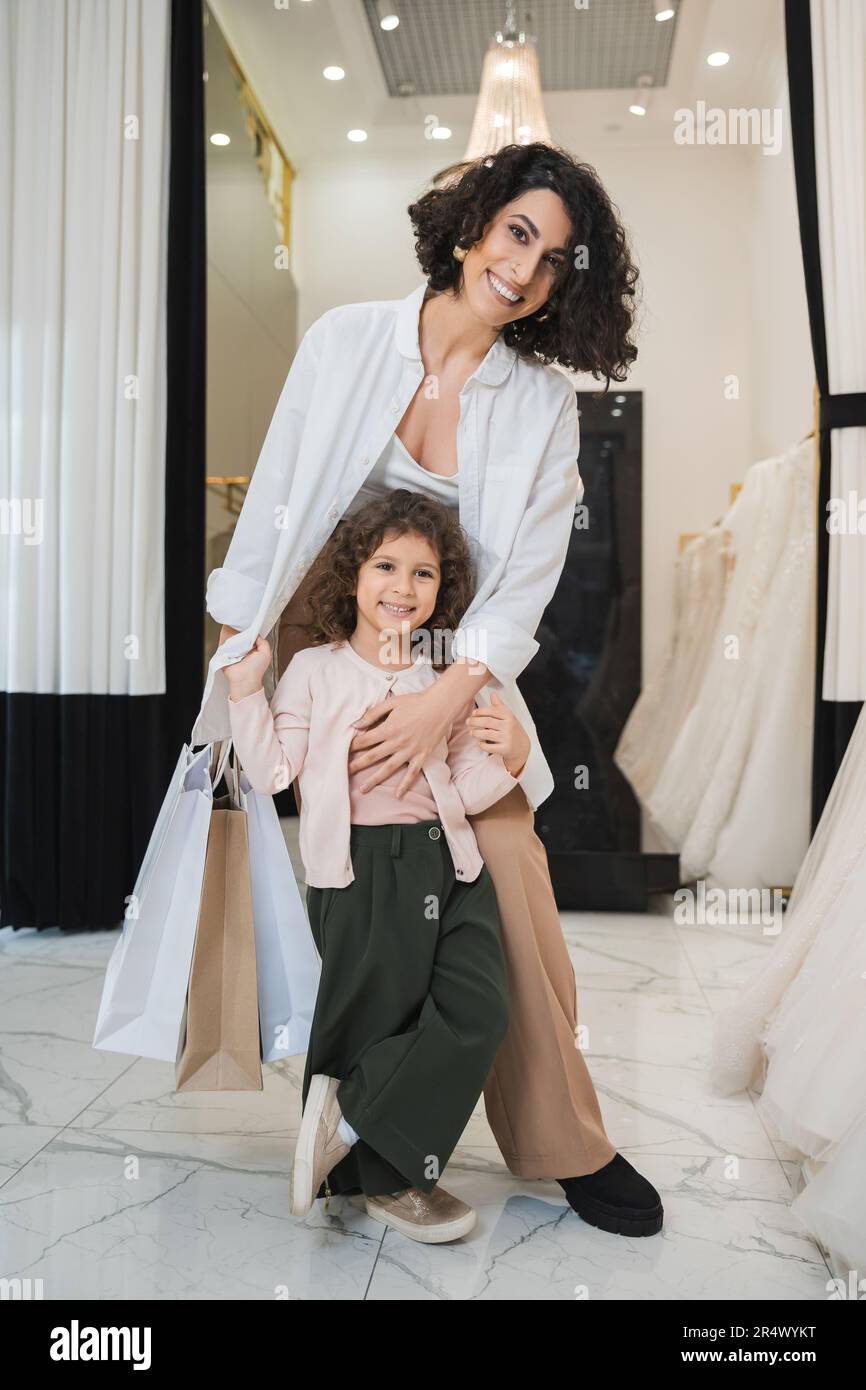 Eine fröhliche Frau aus dem Nahen Osten mit braunem Haar, die Einkaufstaschen in der Hand hält und ein süßes kleines Mädchen umarmt, während sie in Brautkleidern in Brautkleid steht Stockfoto