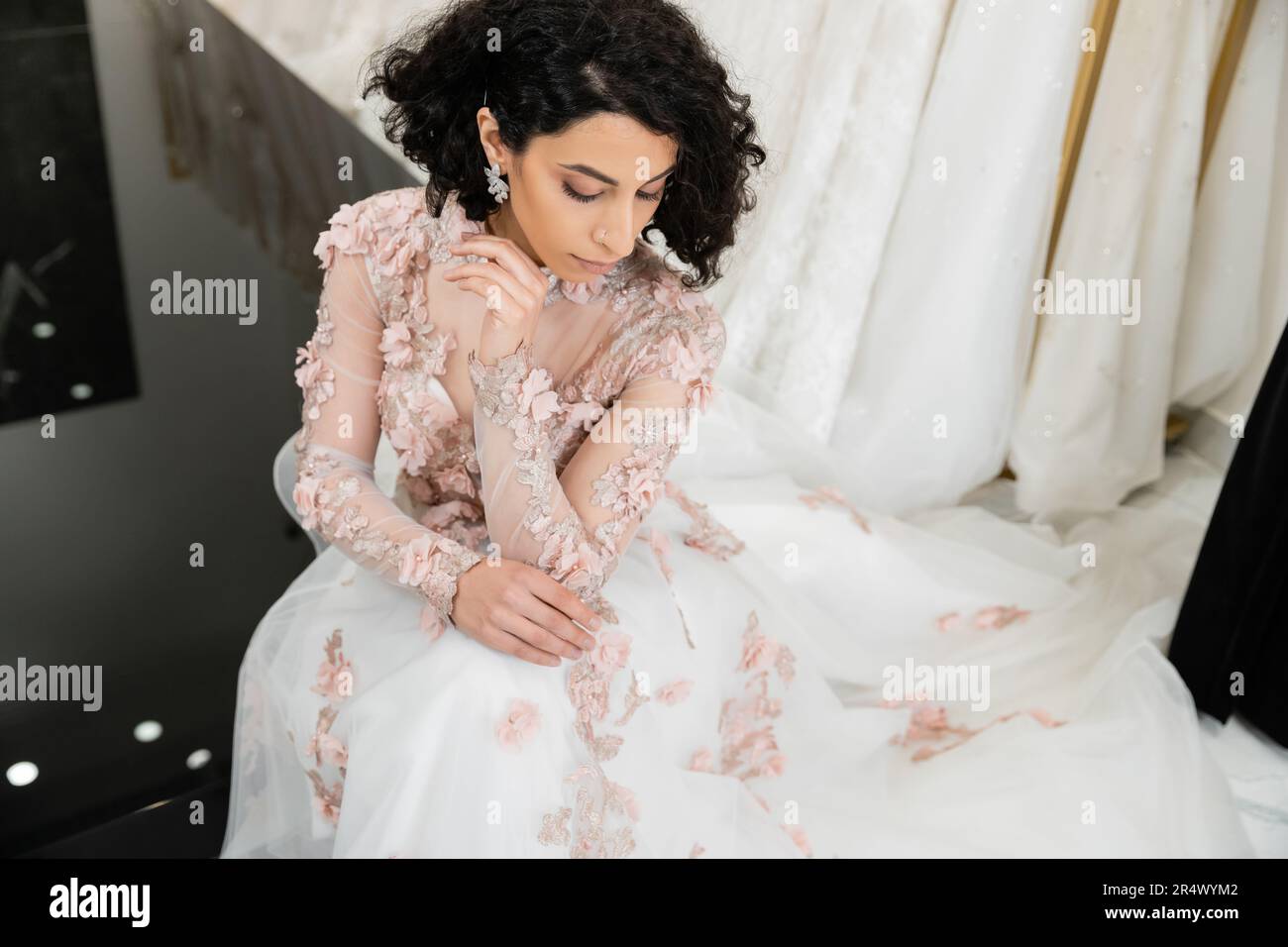 Blick von oben auf eine brünette Frau aus dem Nahen Osten mit welligen Haaren, die in einem wunderschönen und blumigen Hochzeitskleid neben einem verschwommenen und weißen Kleid im Innern des Luxus sitzt Stockfoto