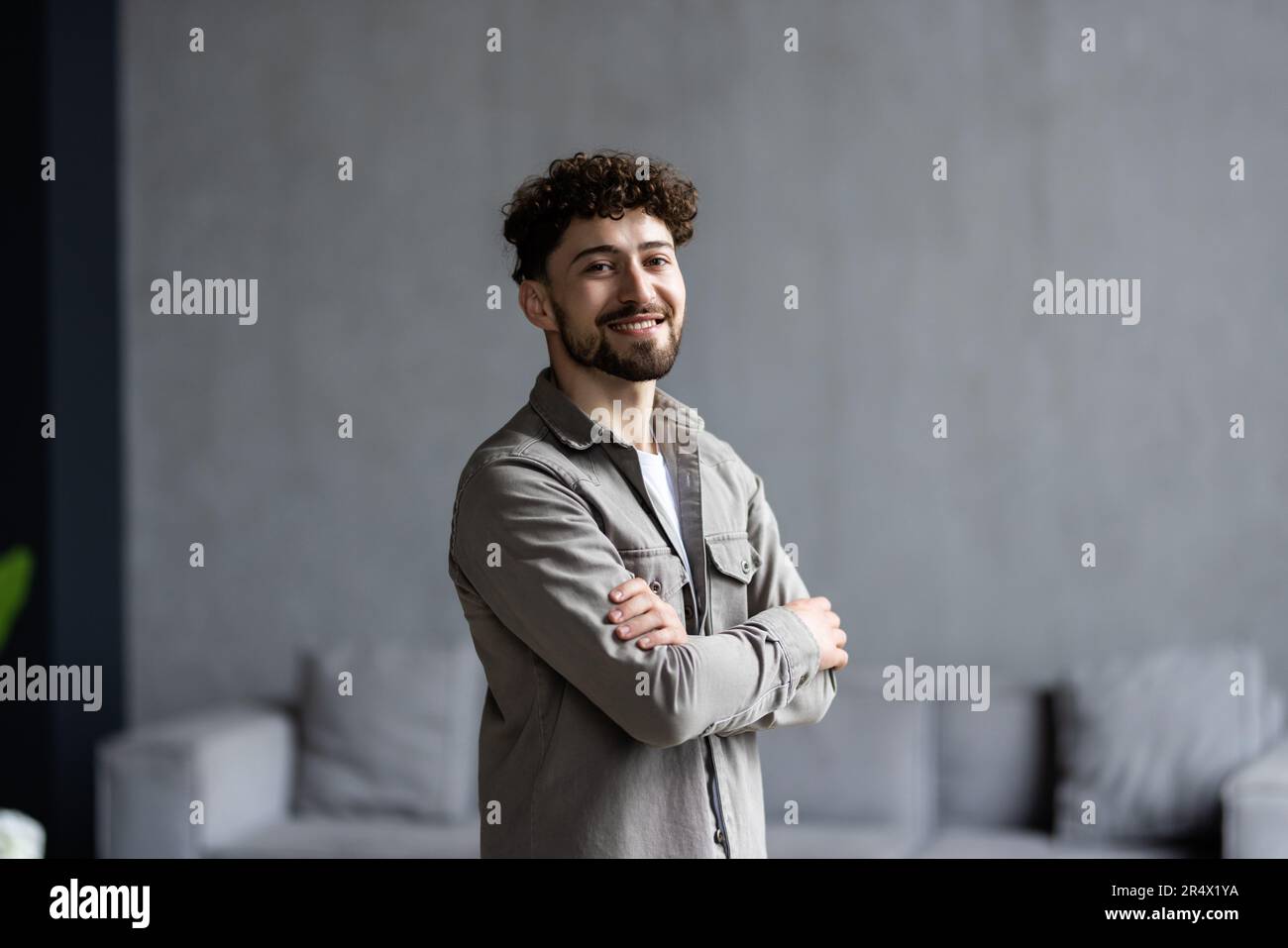 Glücklicher, selbstbewusster Geschäftsmann der Millennials im legeren Stehen mit verschränkten Armen am Arbeitstisch im Heimbüro, der in die Kamera schaut, lächelt. Männlicher Freiberufler Stockfoto