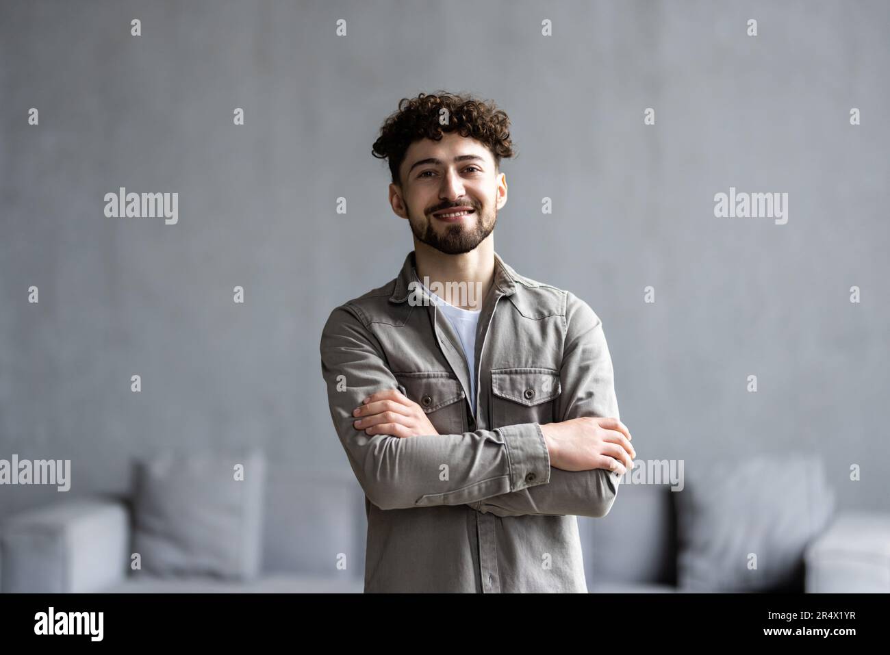 Glücklicher, selbstbewusster Geschäftsmann der Millennials im legeren Stehen mit verschränkten Armen am Arbeitstisch im Heimbüro, der in die Kamera schaut, lächelt. Männlicher Freiberufler Stockfoto