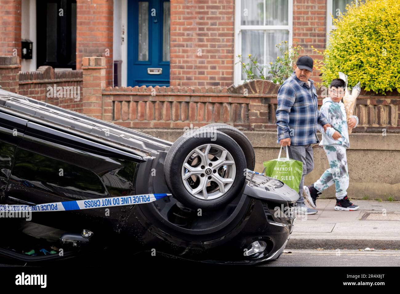 Die Einheimischen gehen am 30. Mai 2023 in London, England, vorbei an den Folgen eines Autounfalls, bei dem ein umgestürzter Nissan-Heck am Straßenrand zwischen Brixton und Camberwell auf der Coldharbour Lane bleibt. Stockfoto
