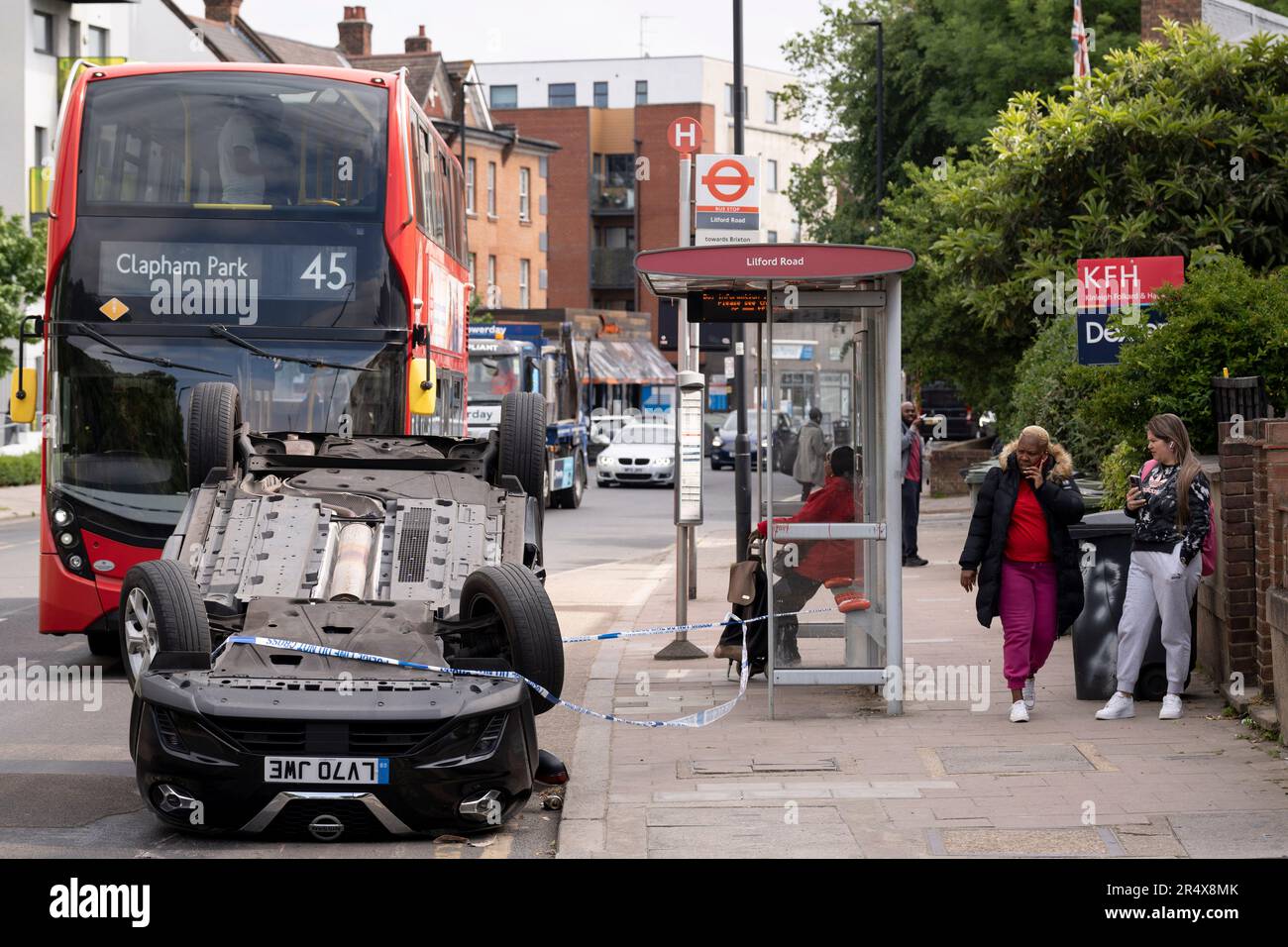 Ein Bus nähert sich den Folgen eines Autounfalls, bei dem am 30. Mai 2023 in London, England, ein umgestürzter Nissan-Heck am Straßenrand zwischen Brixton und Camberwell auf der Coldharbour Lane verbleibt. Stockfoto