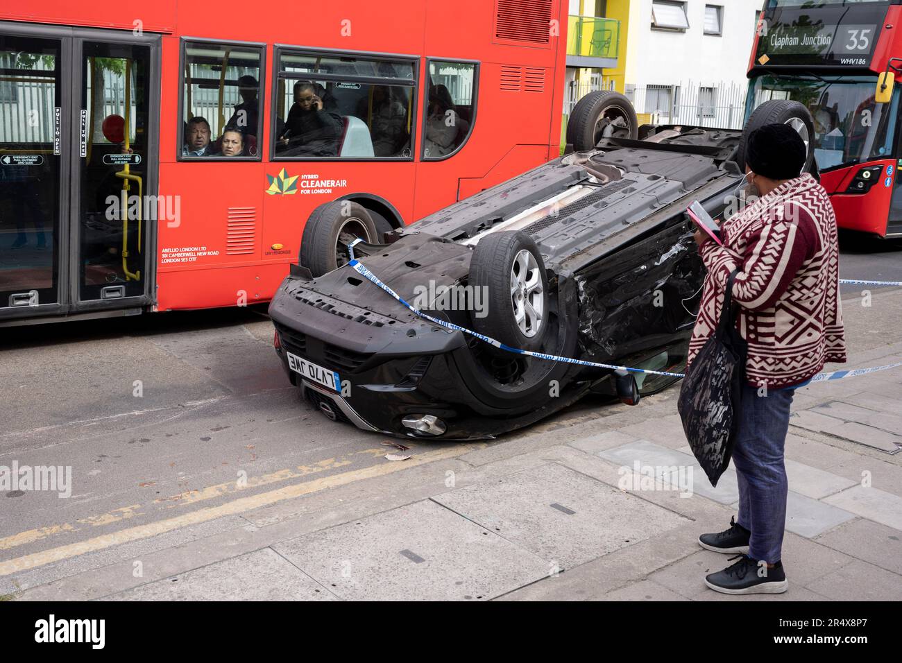Ein Londoner Bus passiert die Folgen eines Autounfalls, bei dem am 30. Mai 2023 in London, England, ein umgestürzter Nissan-Heck am Straßenrand zwischen Brixton und Camberwell auf der Coldharbour Lane verbleibt. Stockfoto