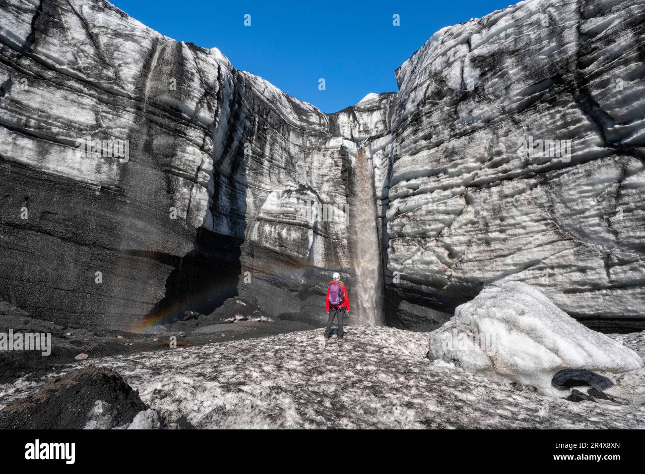 Frau beobachtet einen Wasserfall, der von einer Gletscherwand stürzt, während sie die Landschaft um einen Gletscher im Süden Islands erkundet Stockfoto