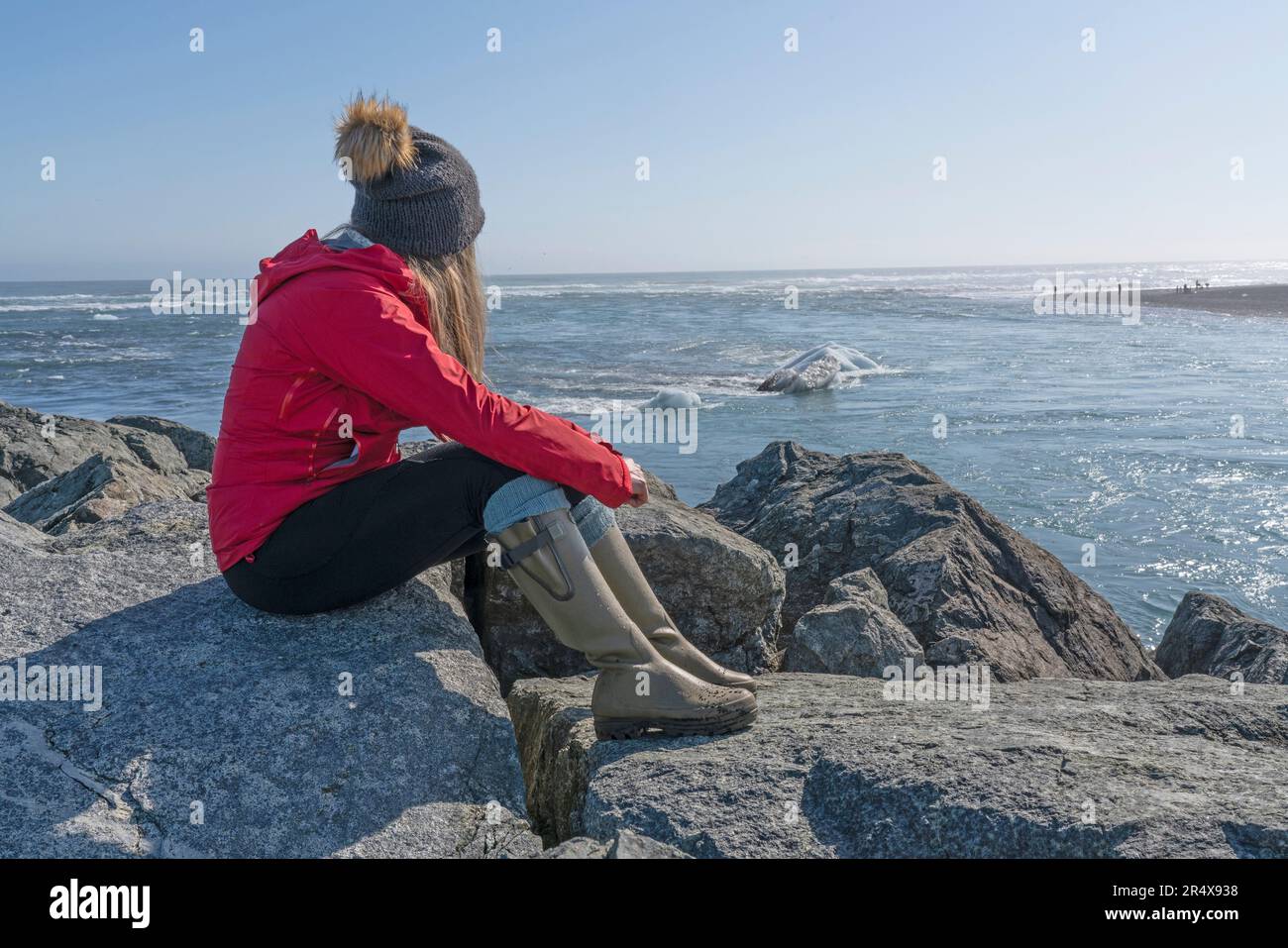 Blick auf eine Frau, die auf den Felsen am Strand sitzt und die Eisberge an der Südküste Islands vorbeiziehen sieht; Jokulsarlon, Süd-Island, Island Stockfoto