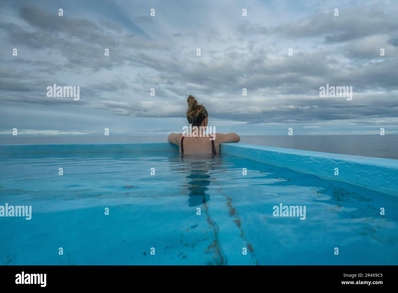 Blick von hinten auf eine Frau, die in einem heißen Pool an der Nordküste Islands sitzt und den Atlantischen Ozean überblickt Stockfoto