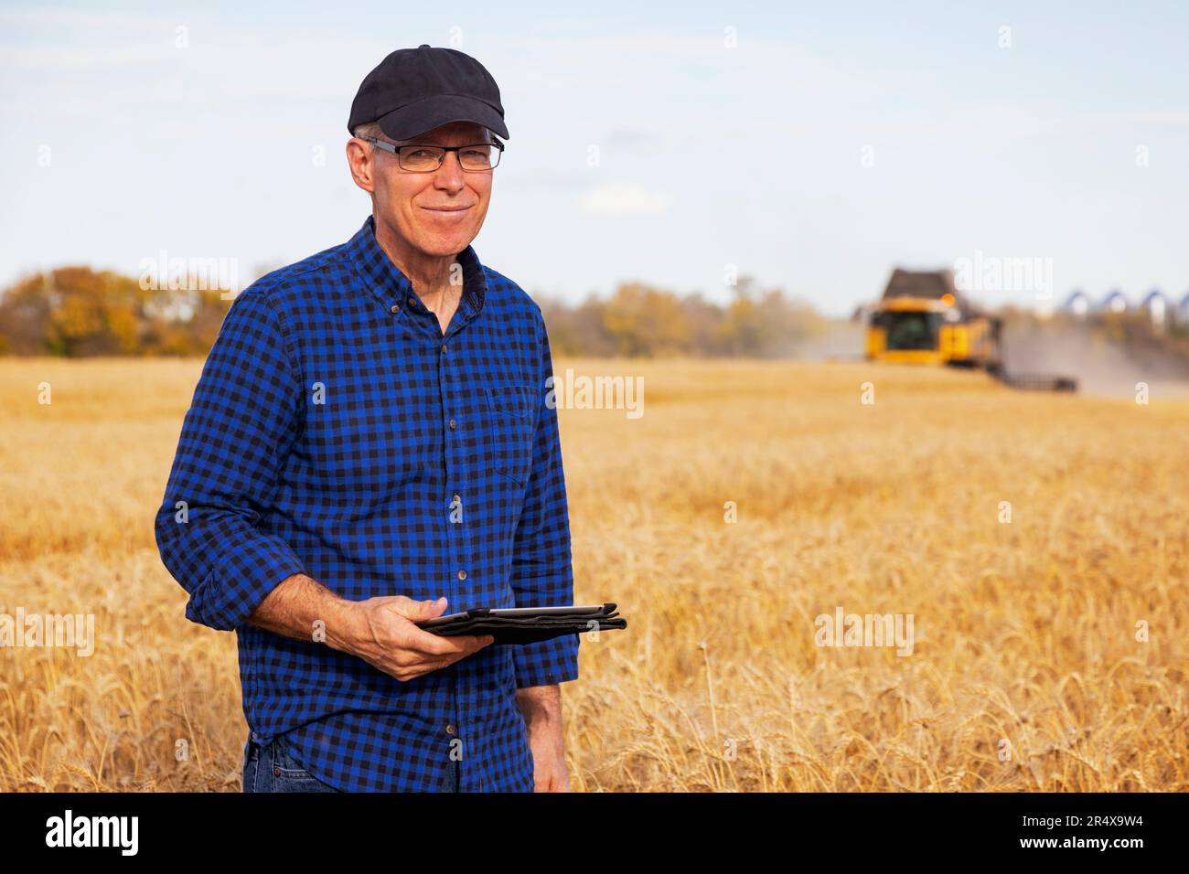 Landwirt, der auf die Kamera blickt, während er mit einem Tablet seine Getreideernte mit Ernteausrüstung im Hintergrund steuert Stockfoto