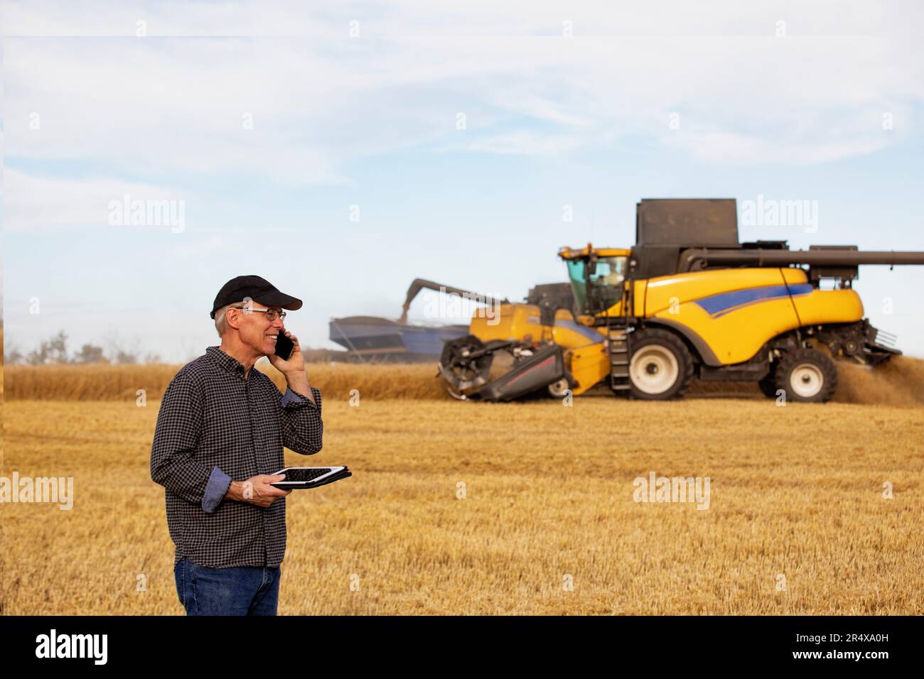 Ein Bauer nutzt ein Tablet, um seine Ernte zu verwalten und spricht mit einem Mähdrescher auf seinem Handy, der im Hintergrund auf einen Getreidewagen abgeladen wird Stockfoto