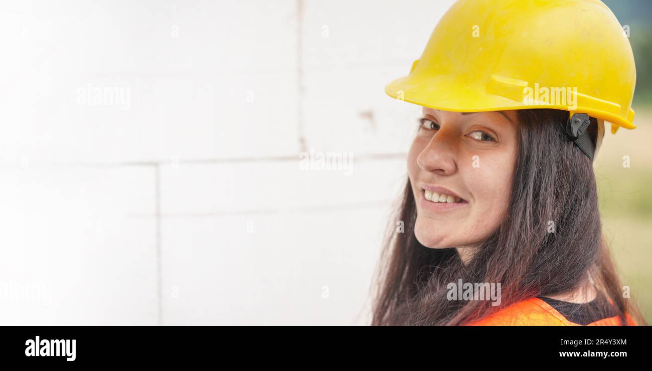 Junge Frau mit gelbem Schutzhelm und orangefarbener Warnweste, langes dunkles Haar, über die Schulter blickend, selbstbewusst lächelnd. Verschwommene Baustelle Stockfoto