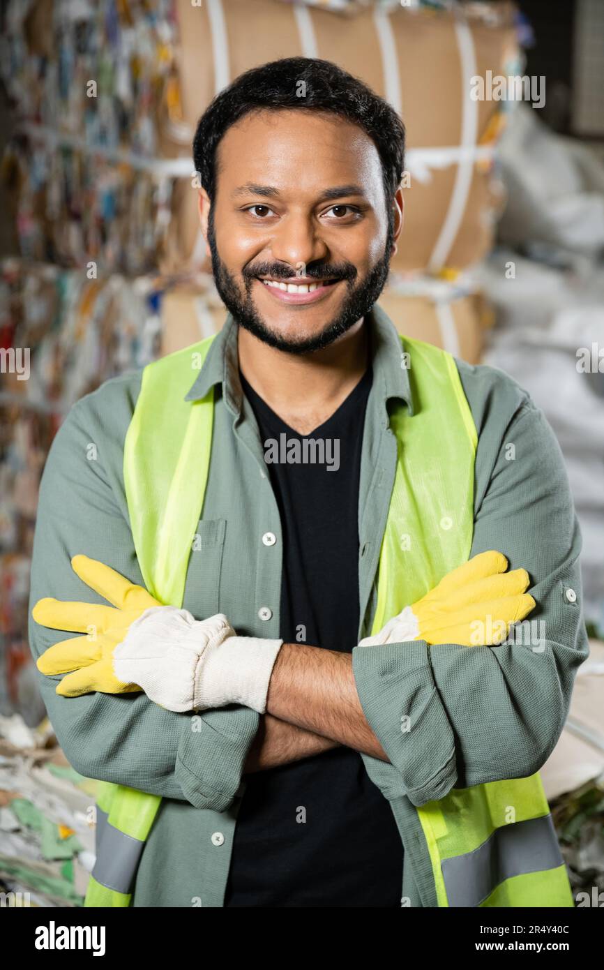 Ein fröhlicher indischer Arbeiter in reflektierender Weste und Handschuhen, der auf die Kamera schaut und die Arme kreuzt, während er neben Altpapier auf verschwommenem Hintergrund steht Stockfoto