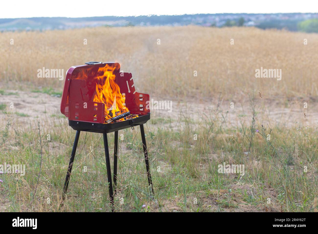 Mobiler Grill oder Barbecue auf einem Boden in der Nähe des Weizenfeldes. Sichtbare Flammen und Feuer auf einem Brennstoff aus Holz und Kohle. Stockfoto