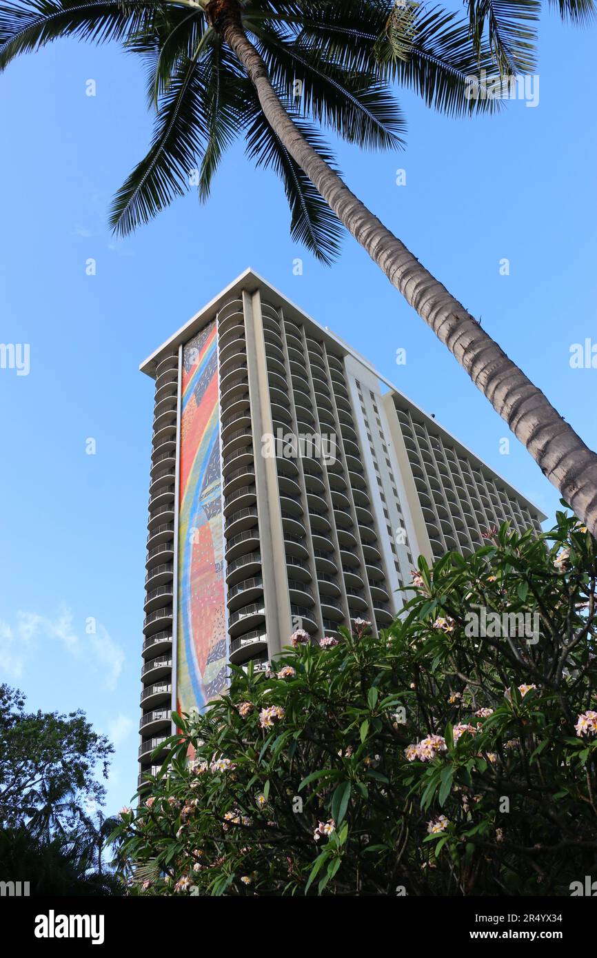 Honolulu, Hawaii, USA - 26. Mai 2023: Vertikaler Blick auf den berühmten Hotelturm des Hilton Hawaiian Village, umrahmt von einer Palme und Plumeria-Blüten. Stockfoto