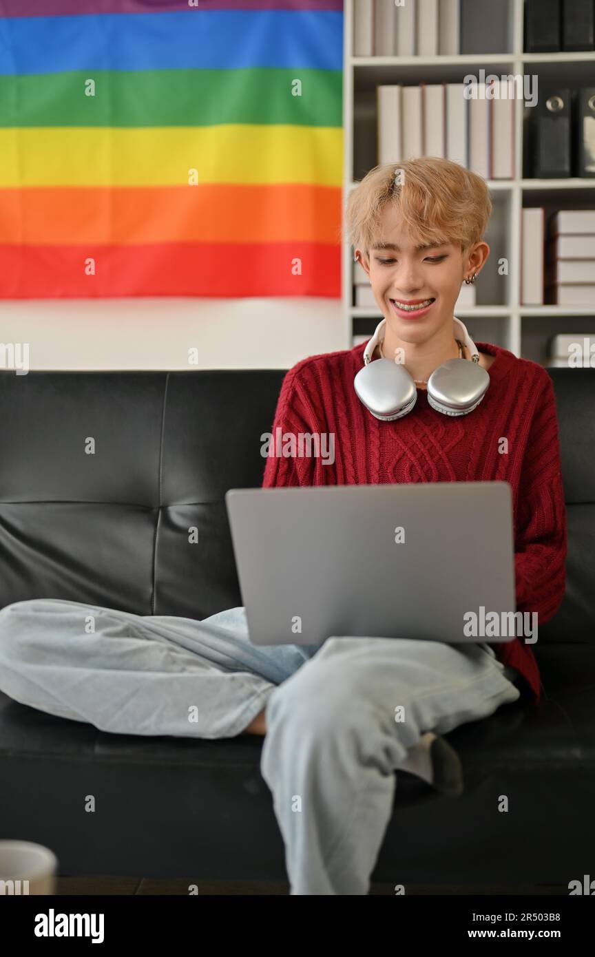 Porträt eines glücklichen jungen asiatischen Schwulen, der seinen Laptop benutzt, während er sich auf einem Sofa in seinem Wohnzimmer entspannt. Stockfoto