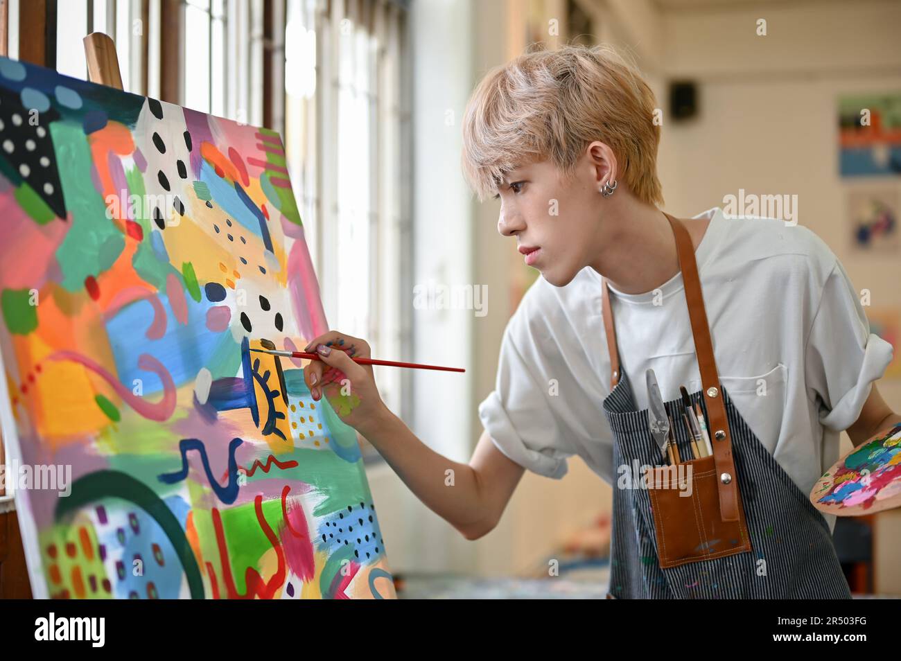 Ein professioneller und talentierter junger asiatischer, schwuler Künstler in einer Schürze konzentriert sich darauf, sein Bild auf einer Leinwand in seinem Studio zu malen. Stockfoto