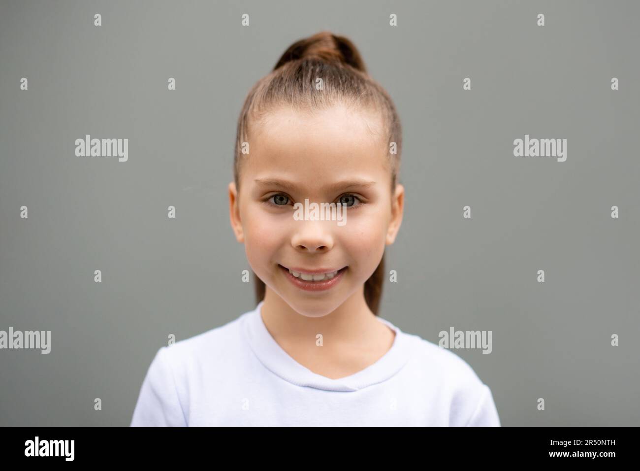 Porträt eines kleinen lächelnden Mädchens in weißem Sweatshirt mit hohem Schwanzhaar auf grauem Hintergrund Stockfoto