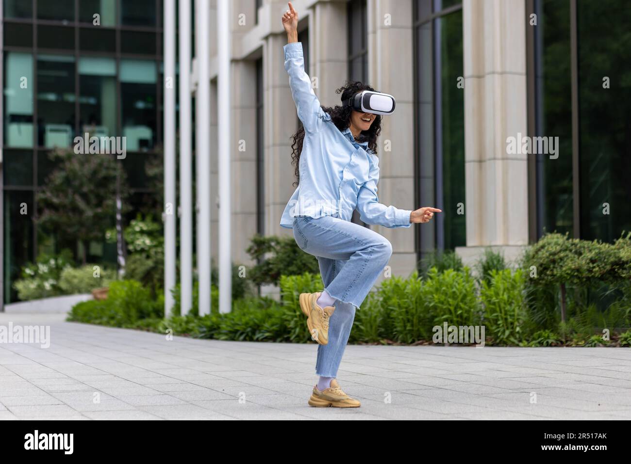 Junge, wunderschöne Frau im Hemd und Jeans, die vor dem Bürogebäude tanzt und eine Virtual-Reality-Brille trägt, eine hispanische Frau, die sich mit lockigem Haar amüsiert und sich entspannt. Stockfoto