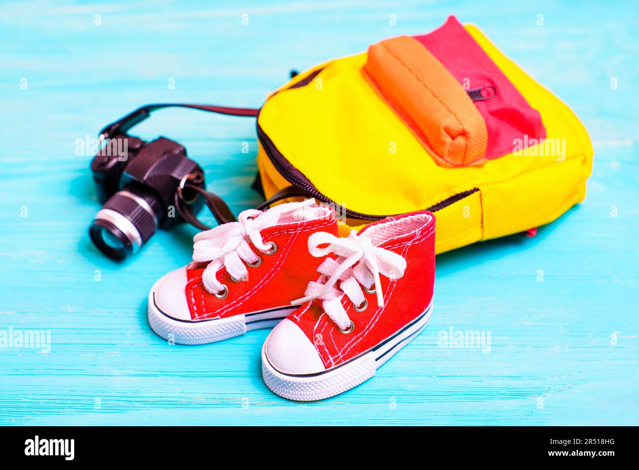 Ein Paar rote Miniaturschuhe, ein kleiner, farbenfroher Rucksack und eine Spielzeugkamera auf blauem Holzhintergrund. Perfekte Kulisse für den Schulanfang oder Stockfoto