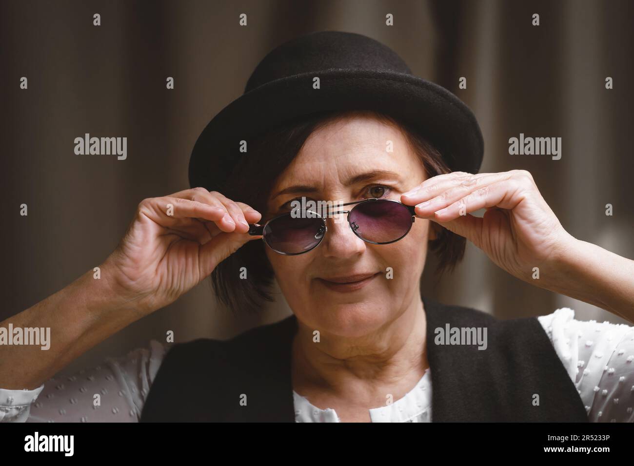 Stilvolles weibliches Modell mittleren Alters mit schwarzem Hut und kurzer dunkler Sonnenbrille, die sich anpasst und im Studio vor dem beigefarbenen Vorhang in die Kamera schaut Stockfoto