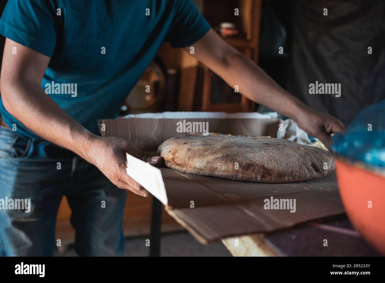Vorderansicht eines gekürzten, anonymen, einheimischen Kochs in blauem Hemd, der am Tisch steht und gebackenes Brot auf einem Karton in der Werkstatt ablegt Stockfoto