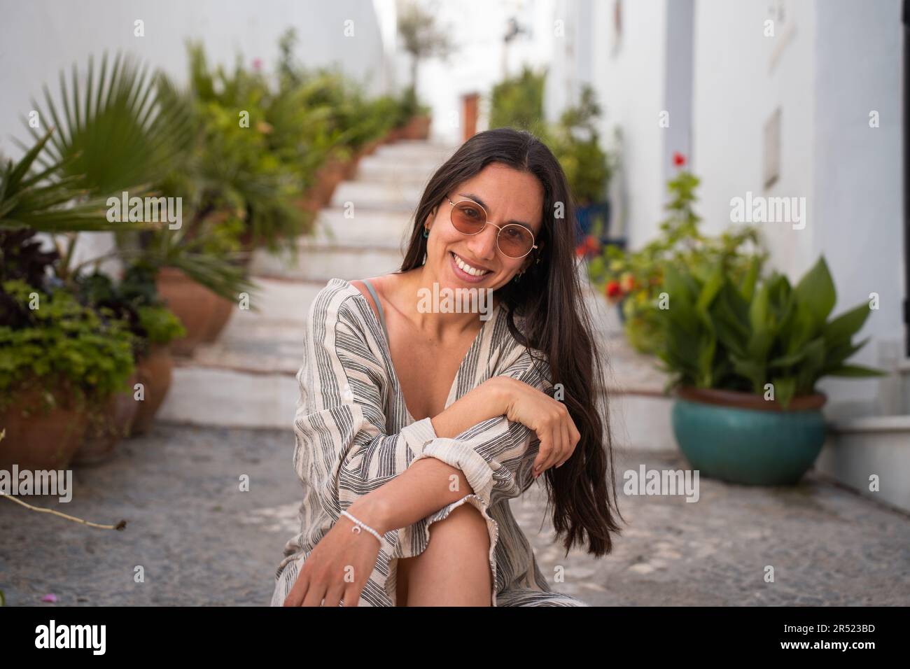 Positive Frau mit Sonnenbrille, die vor der Kamera lächelt. Sie sitzt auf der Straße mit Topfpflanzen vor verschwommenen weißen Gebäuden Stockfoto