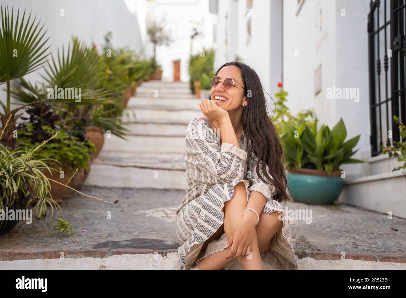 Positive Frau mit Sonnenbrille, die lächelt und mit der Hand am Kinn sitzt, auf der Straße mit Topfpflanzen vor verschwommenen weißen Gebäuden Stockfoto