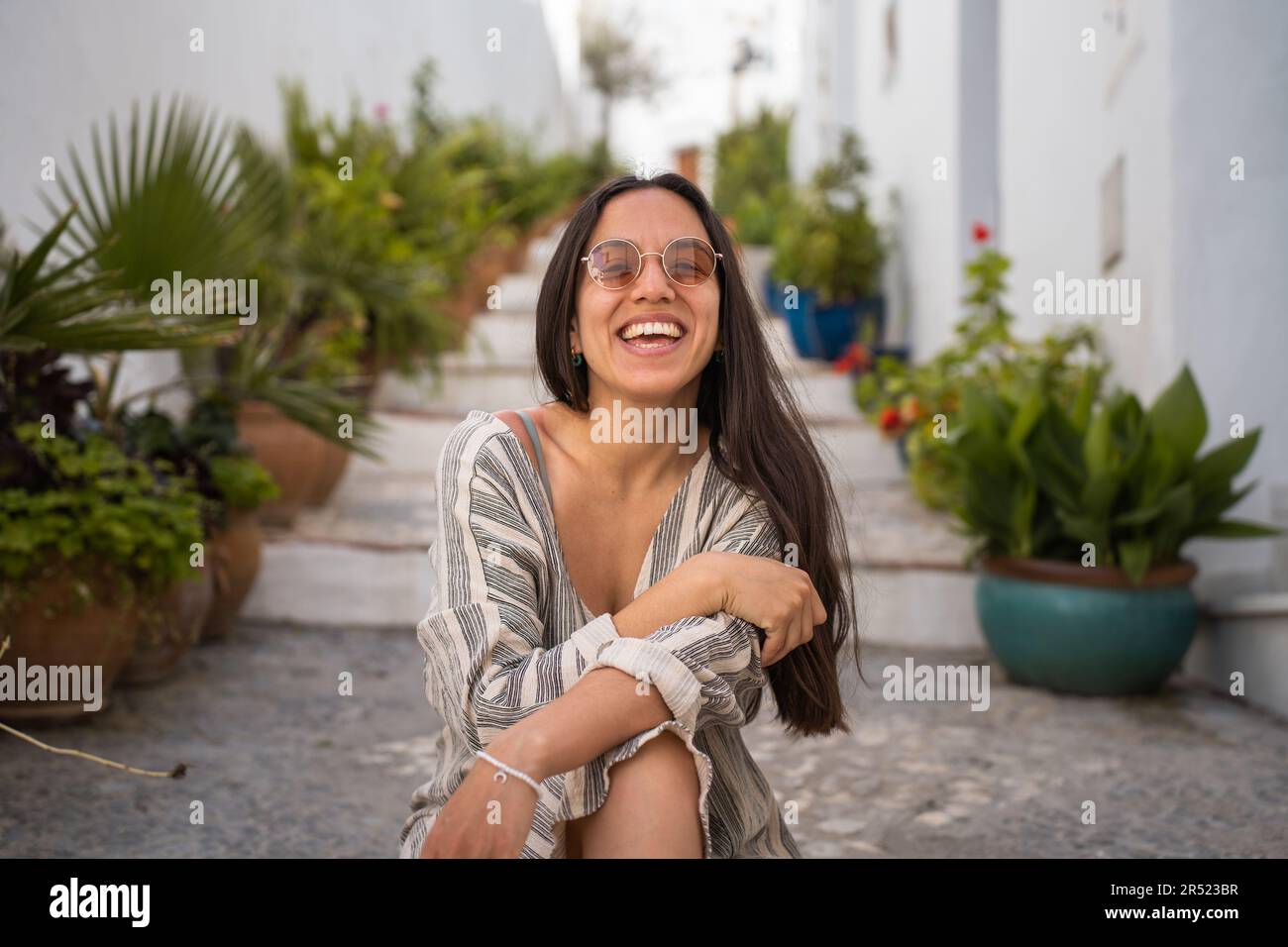Positive Frau mit Sonnenbrille, die vor der Kamera lächelt. Sie sitzt auf der Straße mit Topfpflanzen vor verschwommenen weißen Gebäuden Stockfoto
