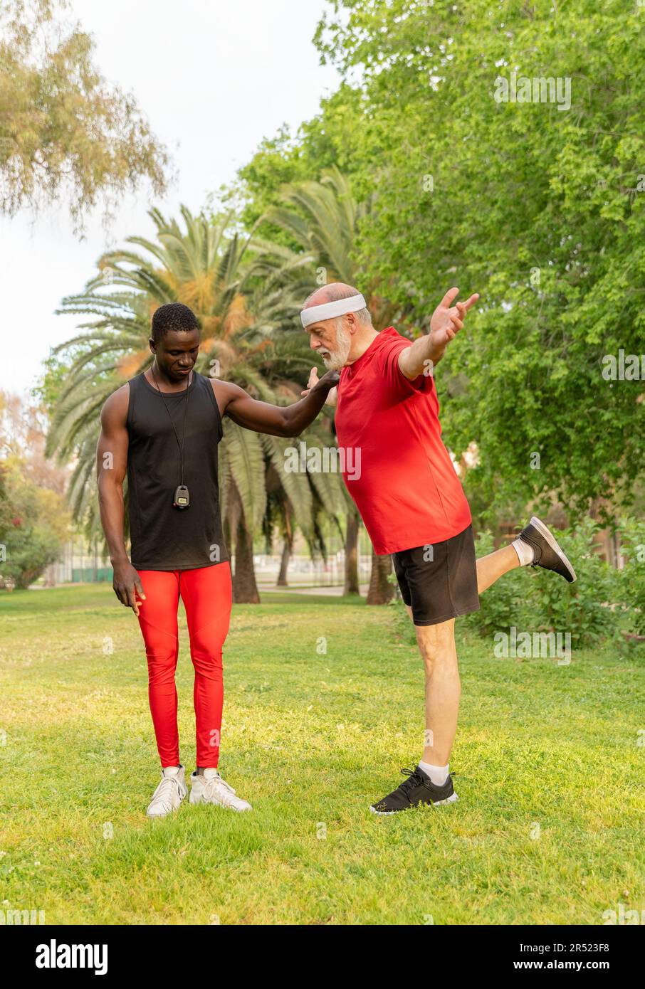 Konzentrierter erfahrener Sportler in Sportbekleidung, der sich vor dem Training Beine streckt, während er mit einem afroamerikanischen Lehrer auf Rasenflächen trainiert Stockfoto