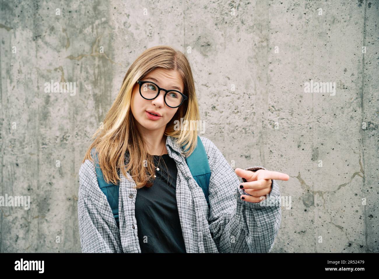 Außenporträt eines jungen Mädchens im Teenageralter mit Brille und Rucksack, Posen auf grauem Hintergrund, Sprechen und Zeigen mit dem Finger zur Seite Stockfoto