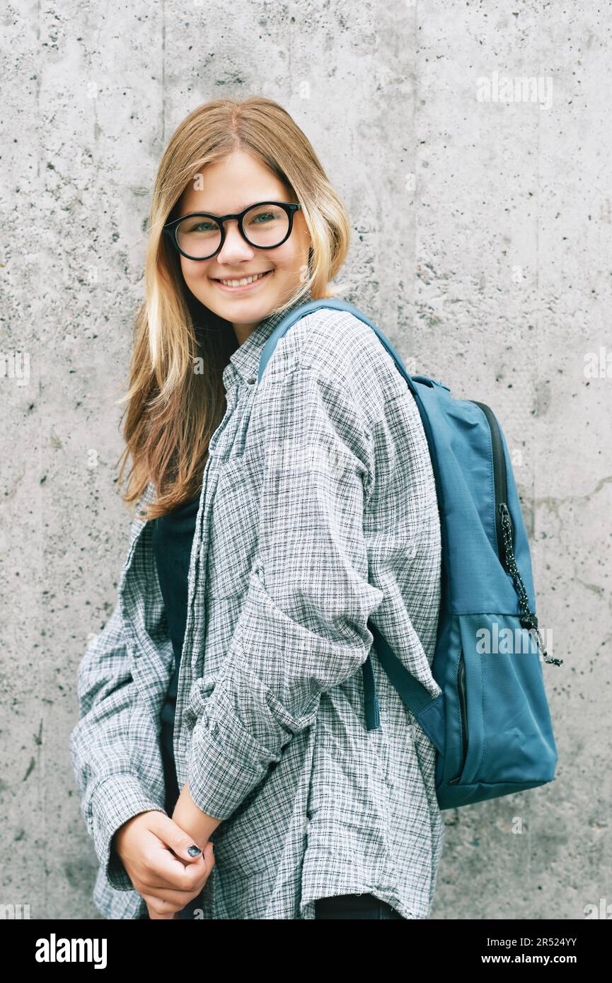 Außenporträt eines jungen Mädchens im Teenageralter mit Brille und Rucksack, das auf grauem Hintergrund posiert Stockfoto