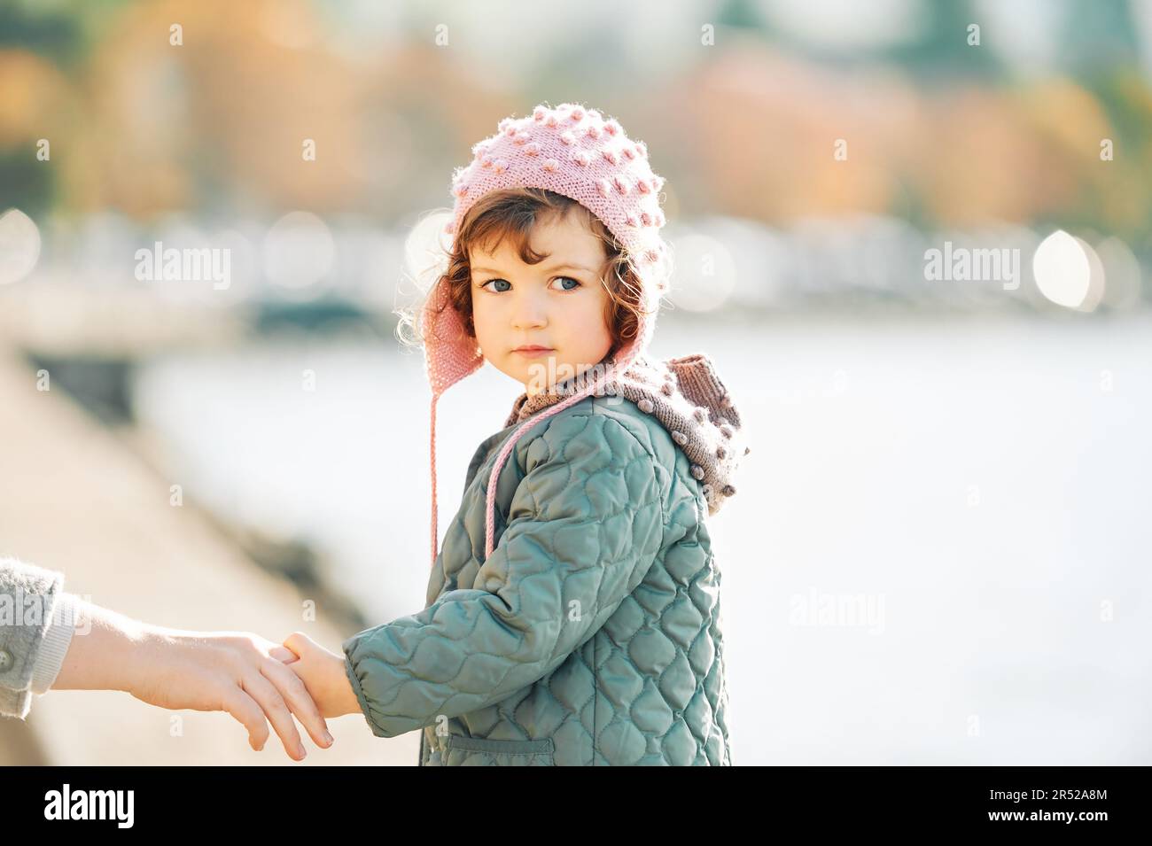 Außenportraot eines niedlichen Kleinkindes, das am See vorbeigeht, moms Hand hält, über die Schulter blickt, eine Jacke mit Aufnäher und einen rosa Hut trägt, kalt Stockfoto