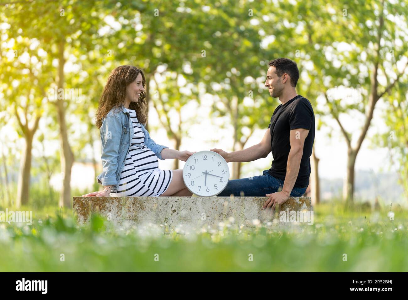Ein Paar mit einer schwangeren Frau, die mit einer Uhr im Park sitzt Stockfoto