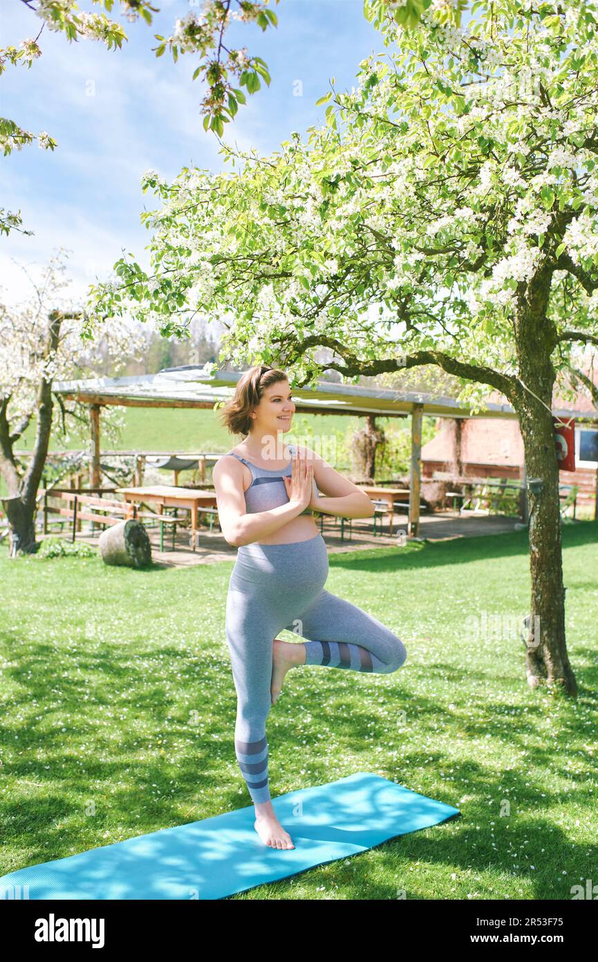 Außenportrait einer glücklichen jungen schwangeren Frau, die Yoga im Frühlingsgarten unter blühenden Apfelbäumen praktiziert, gesunder Lebensstil Stockfoto