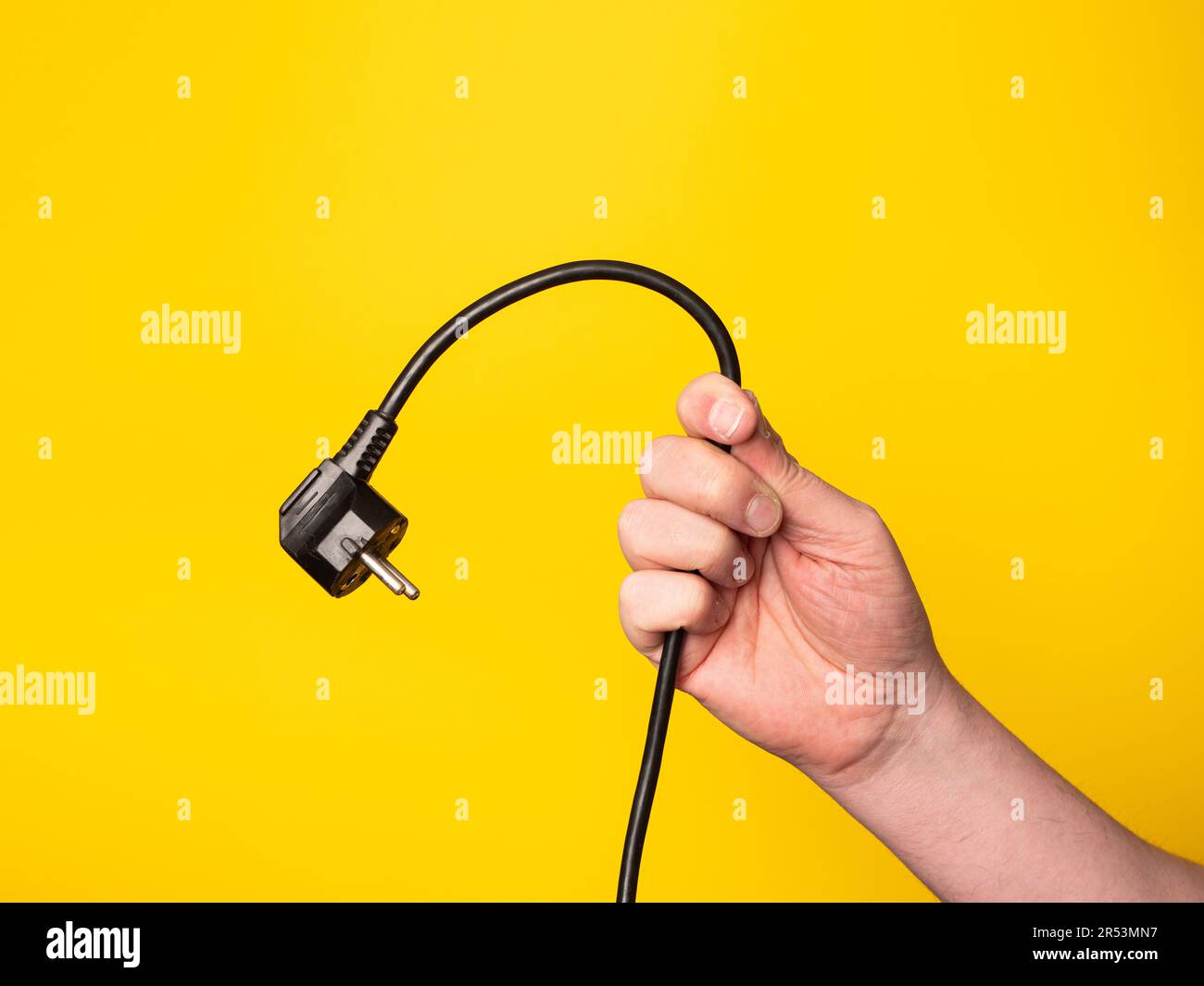 Eine Hand hält einen Stecker, an dem ein schwarzes Kabel befestigt ist. Kein Gesicht, gelber Hintergrund. Stockfoto Eine Hand hält einen Stecker, an dem ein schwarzes Kabel befestigt ist. Kein Gesicht, gelber Hintergrund. Stockfoto