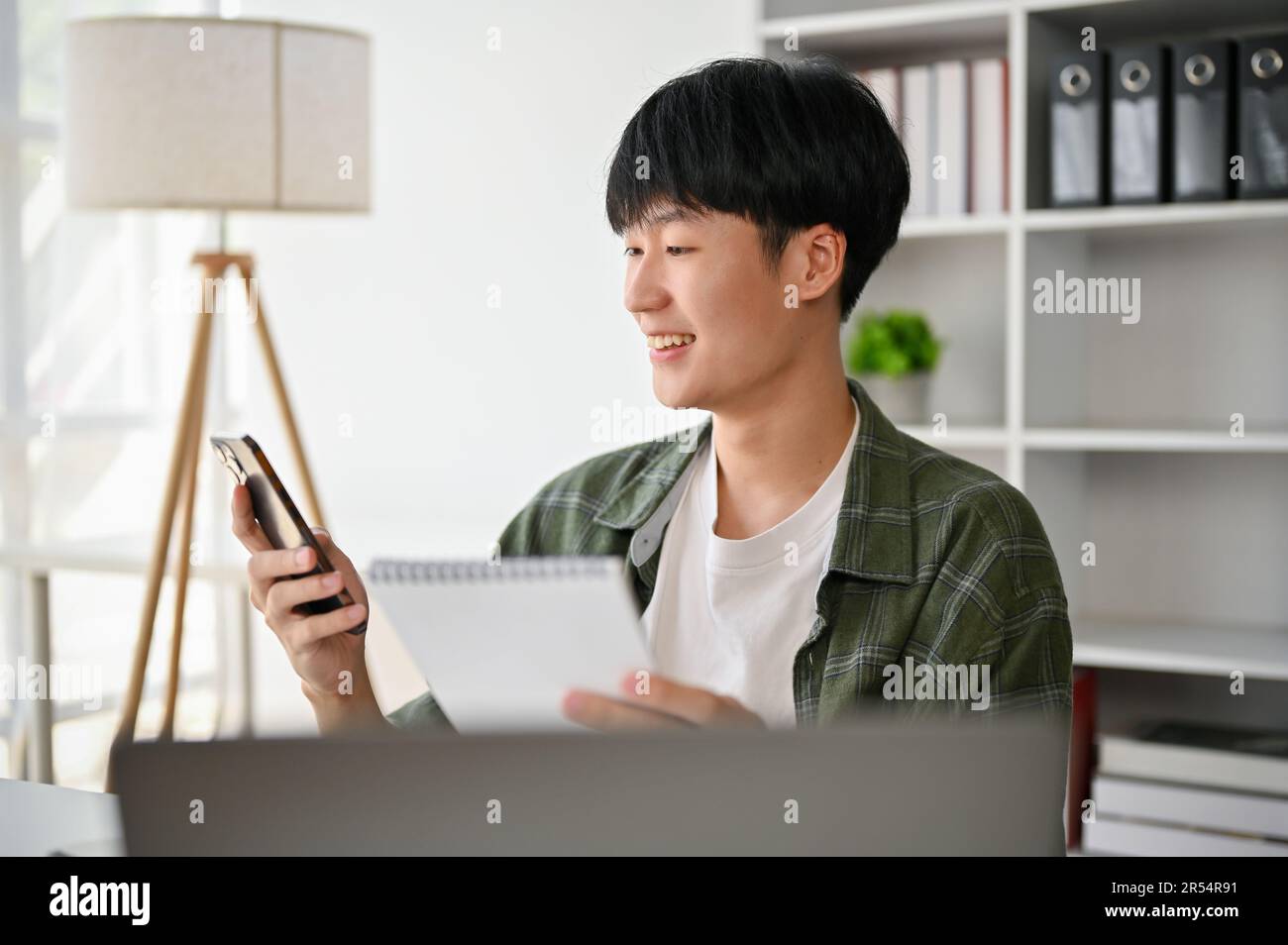 Ein glücklicher junger asiatischer Geschäftsmann oder Marketingassistent tippt am Telefon, sendet Nachrichten oder ruft jemanden an, während er an seinem Schreibtisch sitzt. Stockfoto