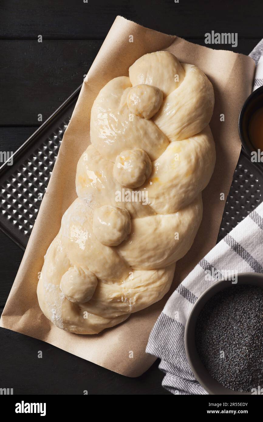 Rohes geflochtenes Brot und Mohnsamen auf schwarzem Holztisch, flach liegend. Traditionelle Sabbat-Challah Stockfoto