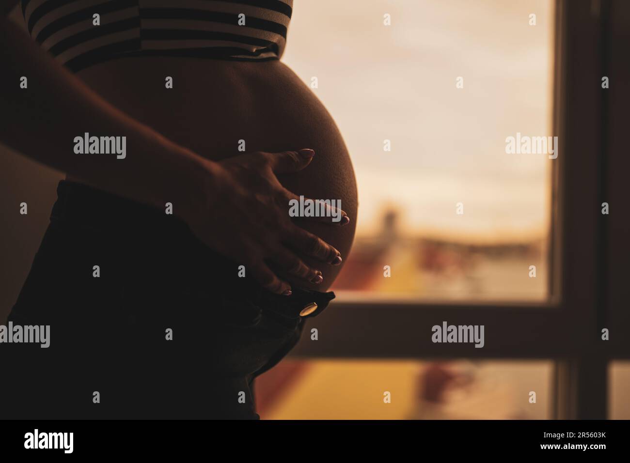 Nahaufnahme einer schwangeren Frau, die neben dem Fenster steht und ihren Bauch hält. Nahaufnahme des Bauch einer schwangeren Frau. Stockfoto