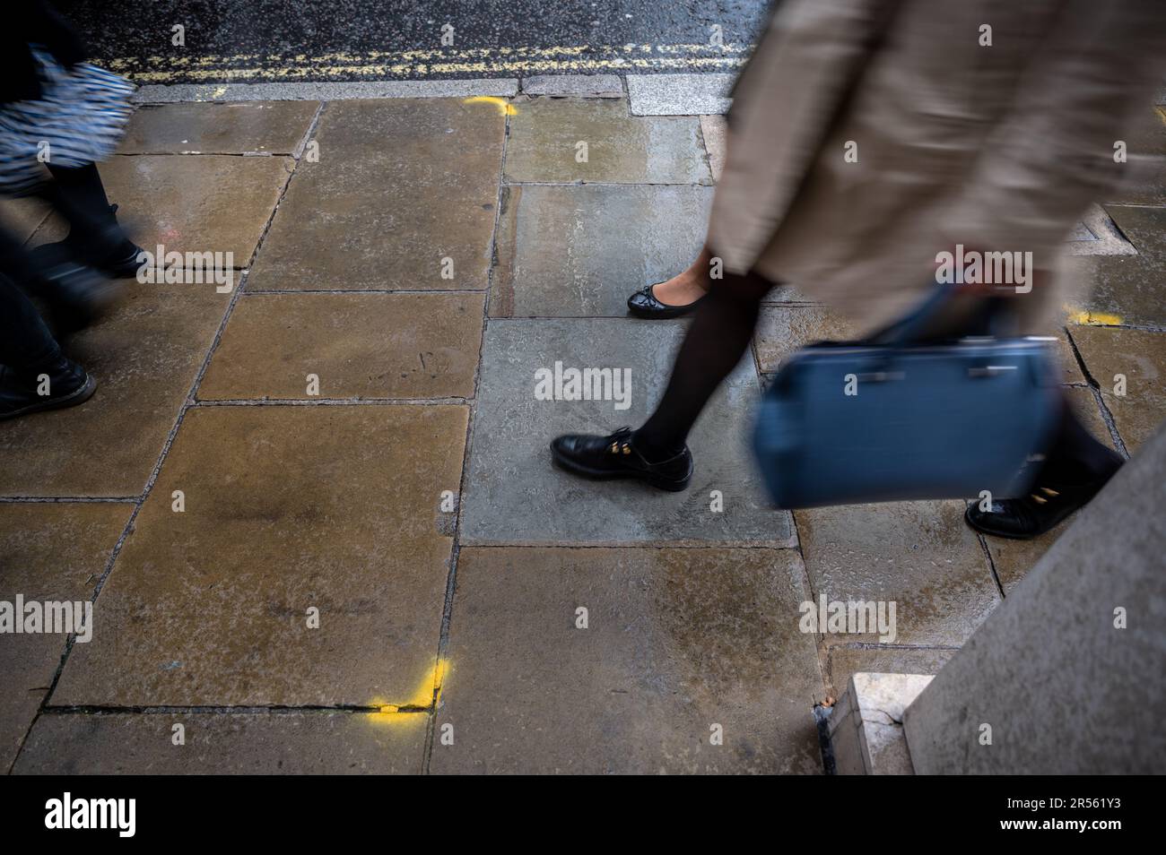 London, Vereinigtes Königreich: Menschen, die im Regen auf einer Straße spazieren. Eine Person trägt eine Handtasche. Ansicht von Beinen und Füßen nur mit Bewegungsunschärfe. Die Stadt London. Stockfoto