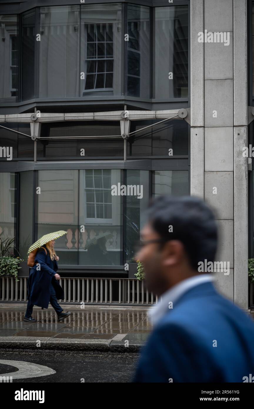 London, Vereinigtes Königreich: Eine Frau läuft im Regen eine Straße entlang und trägt einen Regenschirm. Ein verschwommener Mann ist im Vordergrund und geht in die andere Richtung. Die Stadt London. Stockfoto