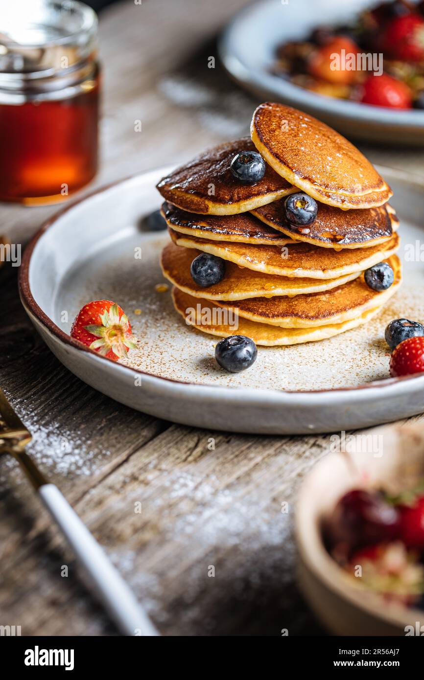 Amerikanische Pfannkuchen mit Heidelbeeren, Erdbeeren und Kirschen mit Honigdip. Auf einem rustikalen Designer-Teller, auf rustikalem Holzbrett. Stockfoto