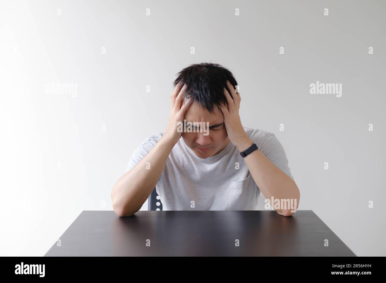 Ein junger Asiate, der ein graues T-Shirt trägt, fühlt sich schwindlig und legt seine beiden Hände auf den schwarzen Tisch. Isolierter weißer Hintergrund. Stockfoto