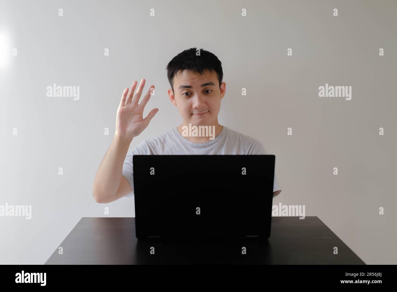 Ein junger asiatischer Mann, der ein graues T-Shirt trägt, winkt mit der Hand zu einem Online-Meeting auf dem Laptop auf einem Holztisch. Isolierter weißer Hintergrund. Stockfoto