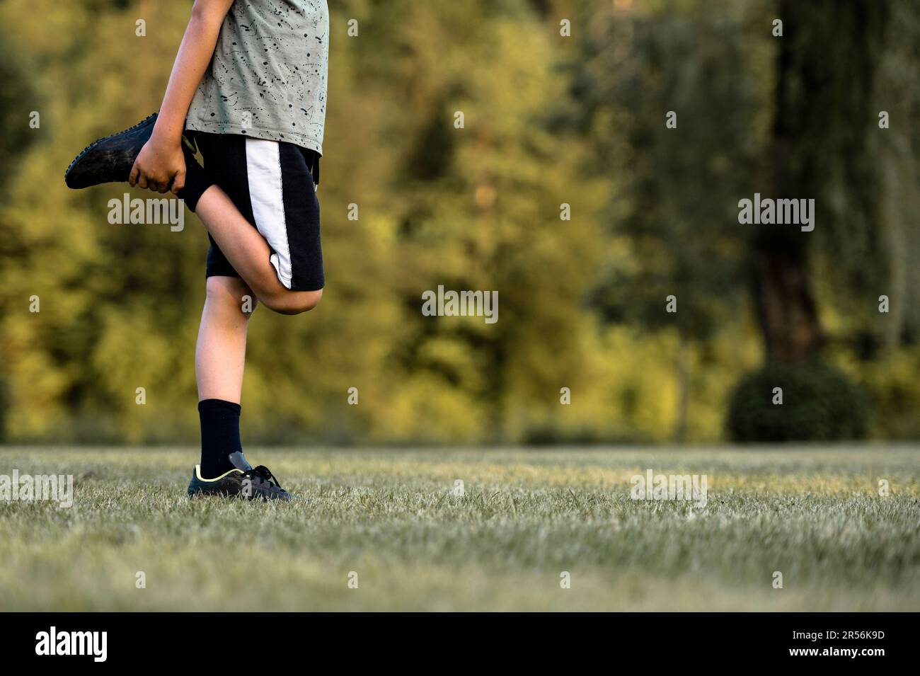 Das Kind wärmt sich vor dem Fußballtraining auf und streckt sein Bein und hält dabei seinen Fuß. Auf einem Spielfeld mit grünem Gras. Stockfoto
