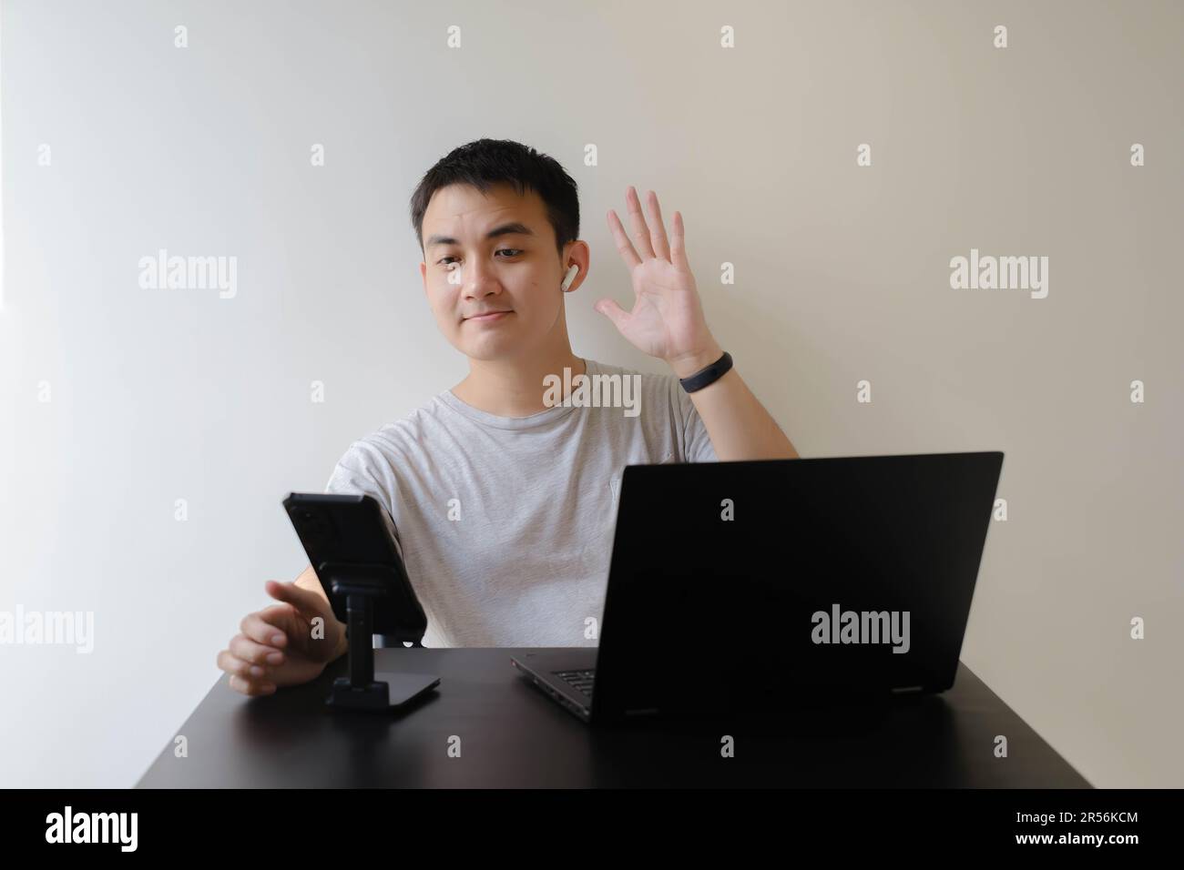 Ein junger asiatischer Mann, der ein graues T-Shirt und ein Paar schnurlose Kopfhörer trägt, winkt mit der Hand bei einem Online-Meeting auf einem Laptop Leuten zu Stockfoto